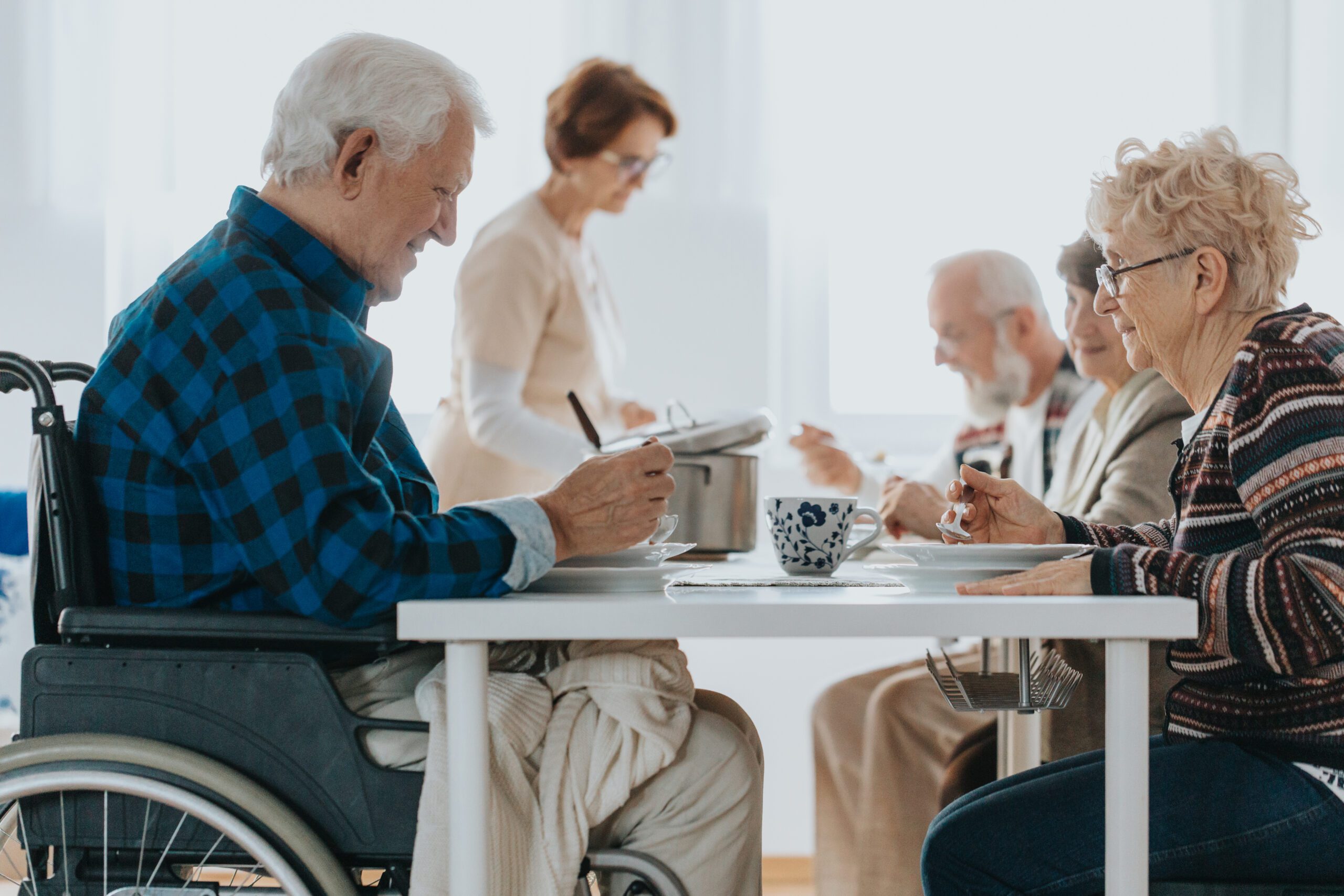 A group of elderly people sitting at a table, enjoying a meal together in a bright, well-lit room; one person is in a wheelchair.