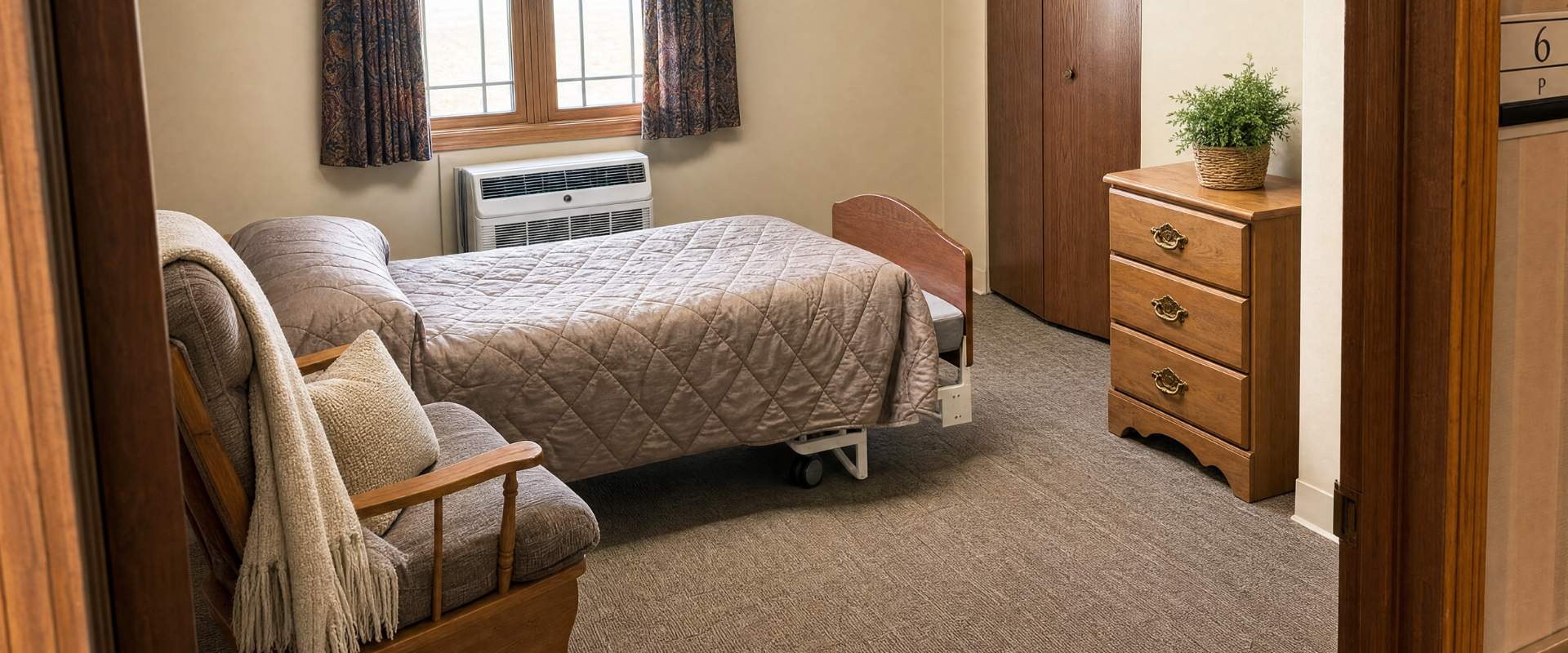 A cozy bedroom with a single bed covered in a quilted beige blanket, a wooden armchair with pillows and a throw blanket, a window with patterned curtains, a wooden dresser with a potted plant on top, and a closed wooden closet door.