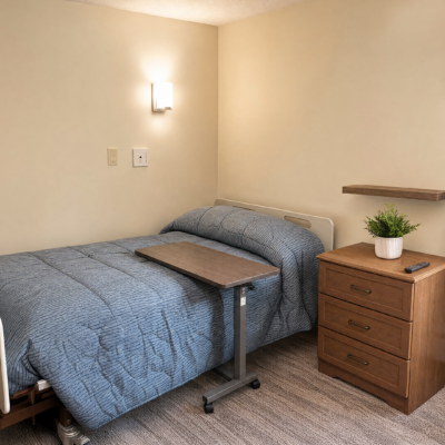 A hospital or care center bedroom with a bed covered in a blue quilt, a wooden bedside table with a potted plant, a wall-mounted light fixture, and a small shelf on the wall.