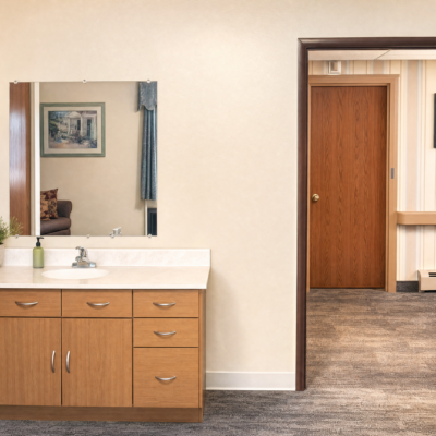 A bathroom vanity with a large mirror and a sink, positioned against a light-colored wall. To the right, an open doorway leads to a hallway with wood-paneled walls and a closed wooden door. The floor is covered with gray carpet.
