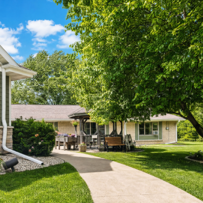 A paved walkway curves through a lush, green lawn with trees and bushes, leading to a light-colored building with large windows and an outdoor seating area under a covered porch. The sky is bright blue with some white clouds.