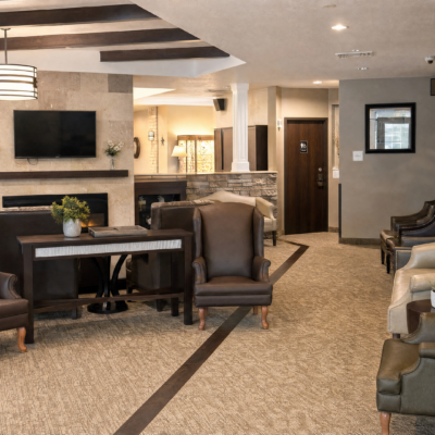 A spacious, well-lit common area with comfortable armchairs and a dining table, featuring a mounted flat-screen TV on a stone accent wall and decorative plants.