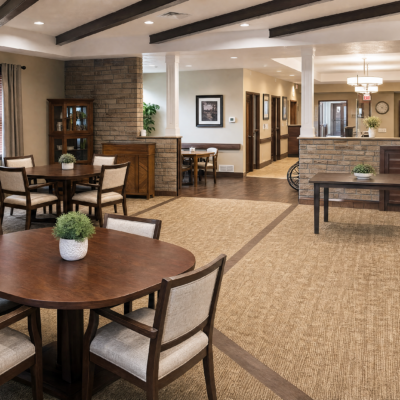 A spacious, well-lit common area with round and rectangular wooden tables, each decorated with small potted plants, and cushioned chairs around the tables. The room features neutral-toned walls, a carpeted floor, and decorative framed artwork, with a partial view of hallway and additional seating areas in the background.
