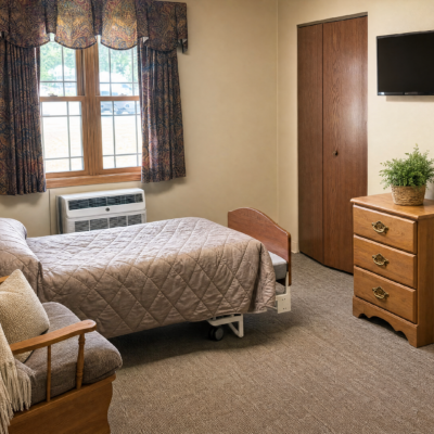 A cozy bedroom with a bed covered in a beige quilt, a window with floral curtains letting in natural light, a wooden dresser with a potted plant on top, a wall-mounted TV, and a small armchair with a blanket draped over it.