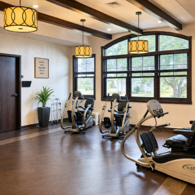A bright, spacious fitness room with large windows overlooking trees, containing several exercise machines, including stationary bikes and a recumbent bike, with hanging yellow pendant lights and a potted plant near the wall.