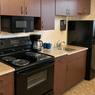 A kitchen area with brown cabinets, a black microwave, a black stove, a coffee maker, and blue mugs on the countertop. There is a stainless steel sink with a faucet, and a black refrigerator to the right.