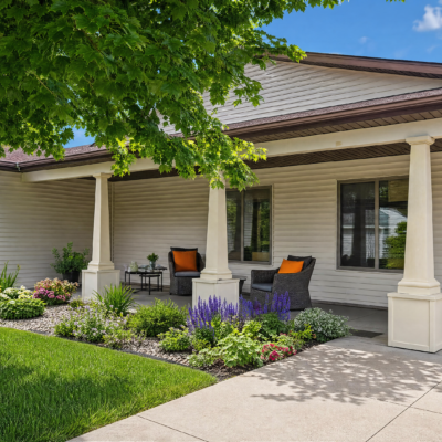 A covered porch with white columns and beige siding, featuring two black armchairs with orange cushions, a small side table, and a garden with various green plants, purple flowers, and a well-maintained lawn.