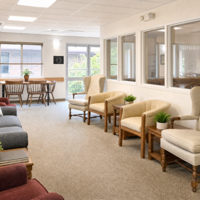 A comfortable lounge area with beige armchairs and small potted plants, large windows allowing natural light, and a dining table with chairs in the background.