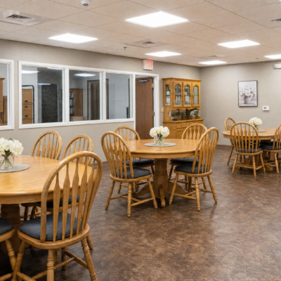 A dining area with round wooden tables, each decorated with white flower arrangements, and surrounded by wooden chairs with black cushions. The room has a large window, a wooden cabinet, and framed artwork on the walls.