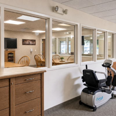 A view of a nursing home activity room seen through large glass windows, with a wooden table and chairs inside, and a stationary exercise bike positioned against the wall outside in a hallway.