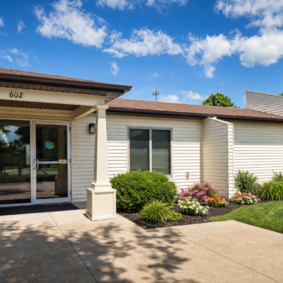 The exterior of a care center building with white siding, a brown roof, and a glass entrance door. There are green bushes and colorful flowers in front, under a bright blue sky with scattered clouds.