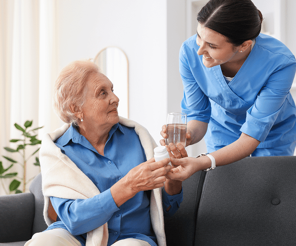 A young female caregiver in blue scrubs offering a glass of water and a pill bottle to an elderly woman sitting on a sofa, smiling warmly at each other in a bright, comfortable room.