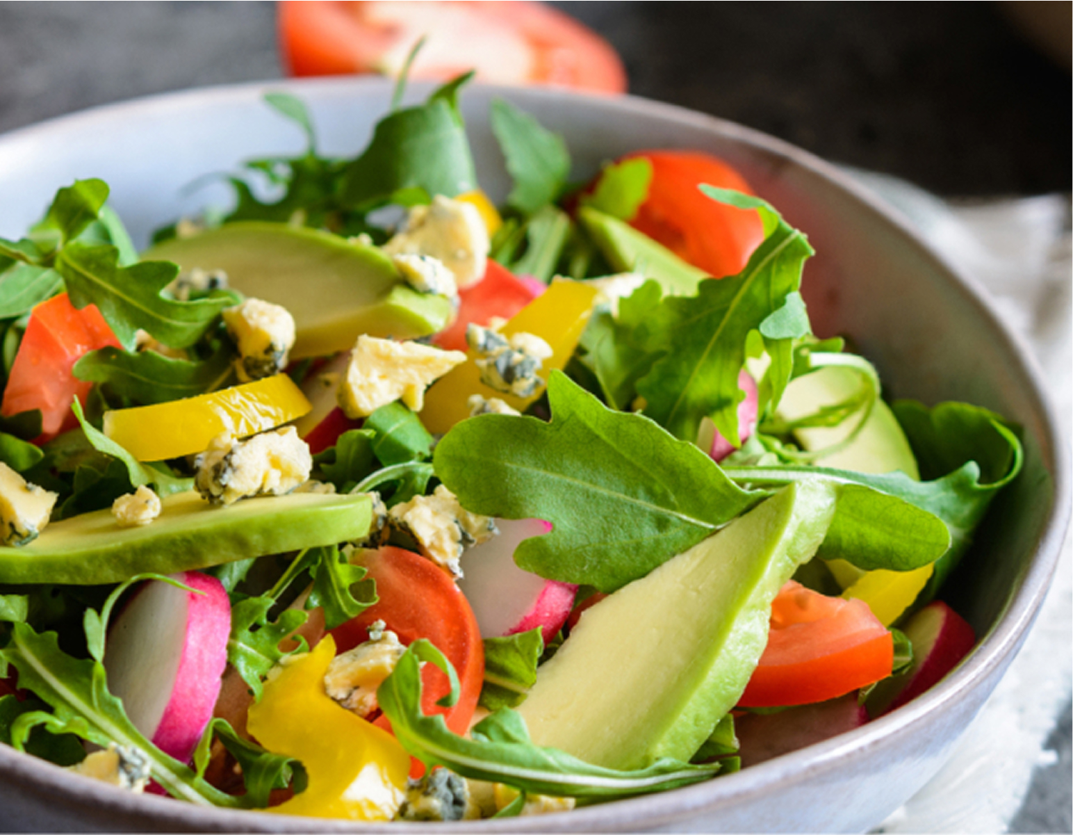 A bowl of fresh salad with sliced tomatoes, avocado, yellow bell peppers, radishes, leafy greens, and blue cheese crumbles.