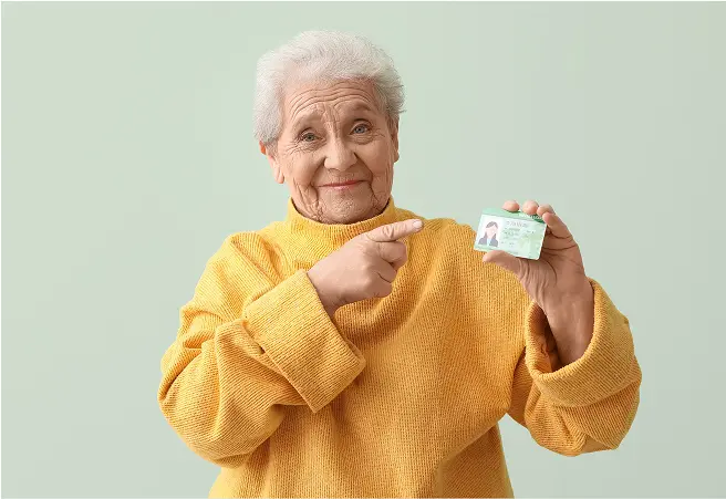 An elderly woman with gray hair, wearing a yellow sweater, holding a driver's license in her right hand, and pointing at it with her left index finger, smiling gently against a light green background.