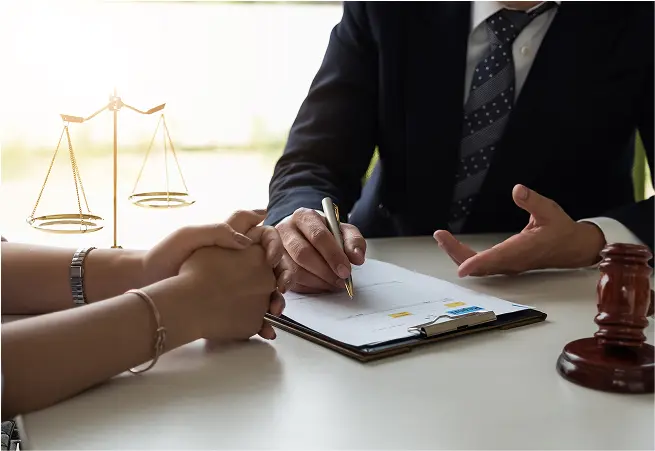 A person in a suit and tie is sitting at a desk, signing a document with a gold pen, while another person, whose hands are visible, holds their hands in support. On the desk, there is a wooden gavel, a document on a clipboard, and a small scale of justice in the background.
