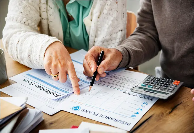 Two people are sitting at a table reviewing insurance documents. One person is pointing at a section of the paperwork while the other is holding a pen ready to sign. There is a calculator on the table, along with stacks of papers and folders.