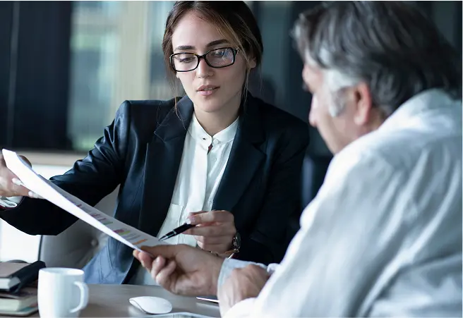 A young woman in a business suit and glasses discusses documents with an older man in a white shirt at a table in a modern office setting.