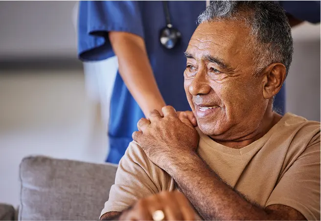 A senior man smiling as a healthcare professional in blue scrubs stands behind him, with a stethoscope around their neck, in a clinical setting.