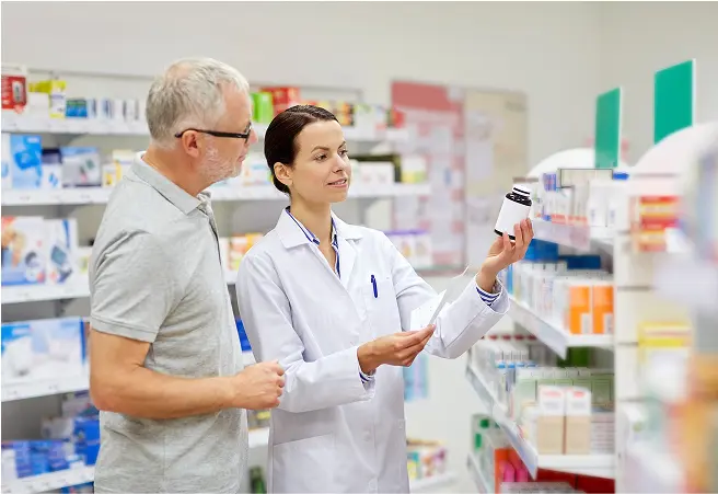 A pharmacy technician shows a medication bottle to an older male customer in a pharmacy aisle, with shelves of medication boxes behind them.